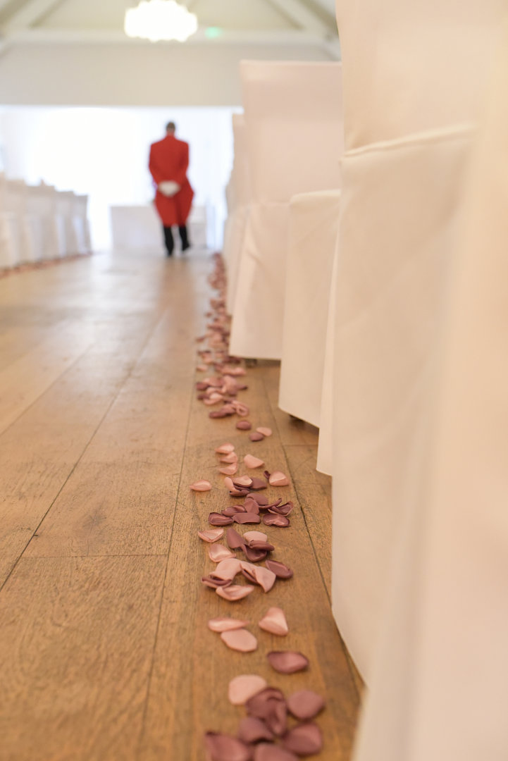 a row of pink and red rose petals down the aisle of a wedding ceremony room with white chairs, toastmaster in his red jacket in the background