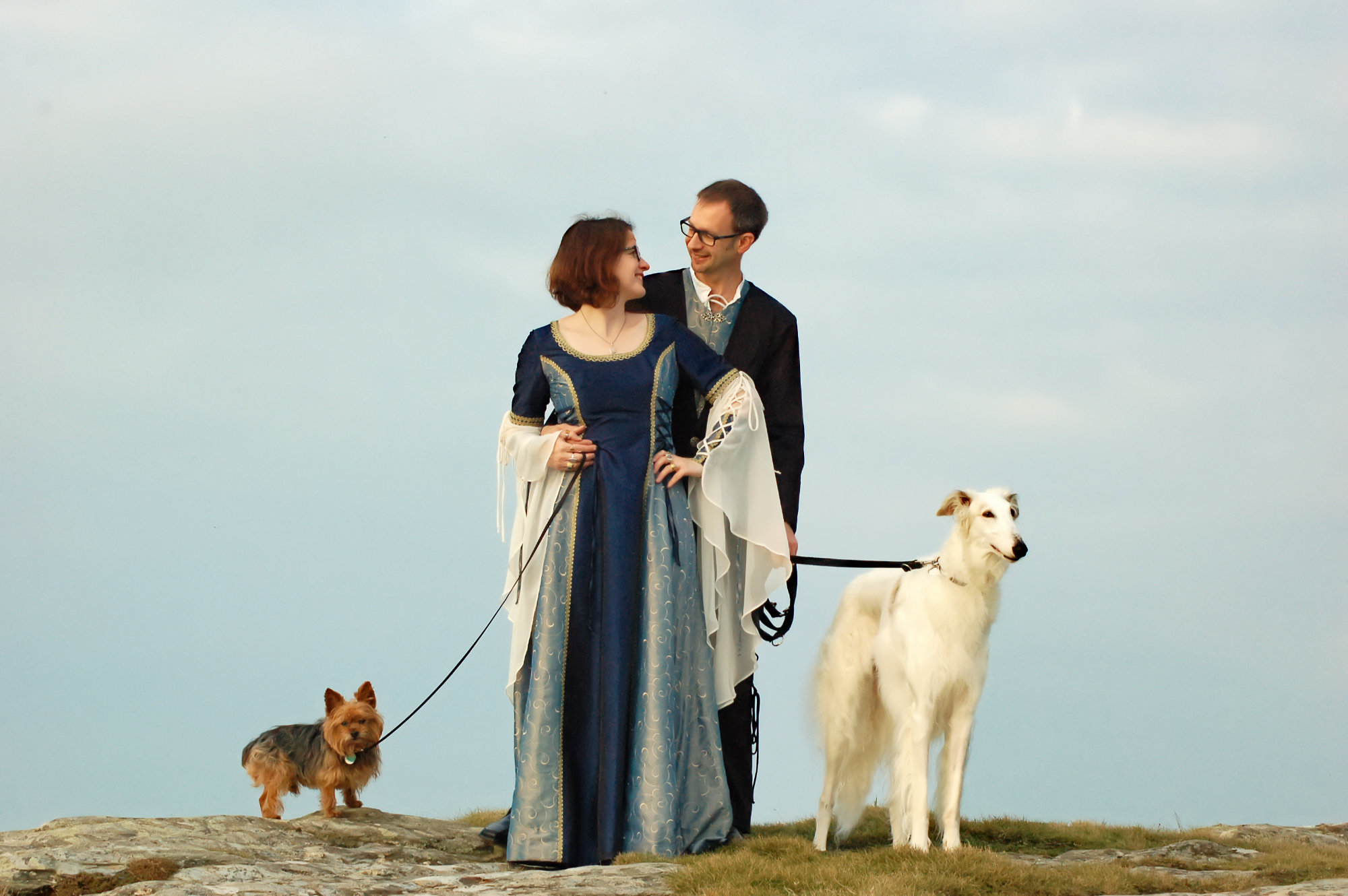 Logo elements ARROWS copyright Nadina Bee Photo of an alternative bride and groom on a cliff with two dogs: a Yorkshire terrier and a white borzoi. The bride wears a medieval style wedding dress in shades of blue, with ivory bell sleeves and gold braiding. The groom wears a black suit with a long jacket. Photo for blog should you take the dog on holiday